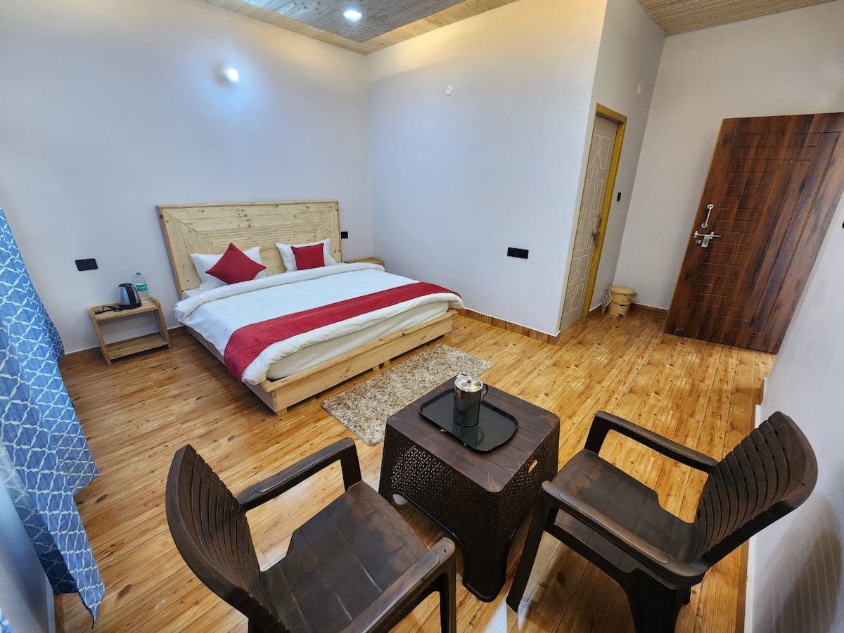 A comfortable room features a spacious bed with a red and white duvet, flanked by two decorative pillows. A small table with a candle sits between two dark chairs. Natural wood flooring and wall accents create a warm atmosphere, complemented by soft lighting and a partially open door.