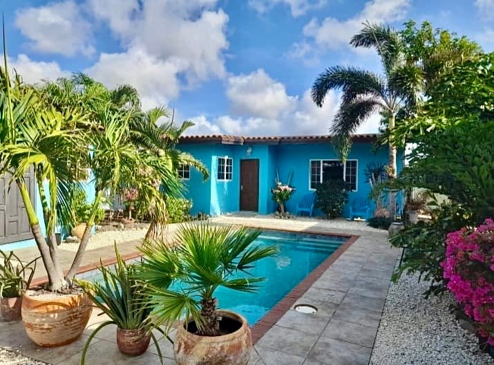 A vibrant blue guest house is seen surrounded by lush palm trees and flowering plants. A sparkling private pool is positioned in the foreground, with sunlit tile areas leading to the entrance door. The bright sky complements the tropical ambiance of the space.