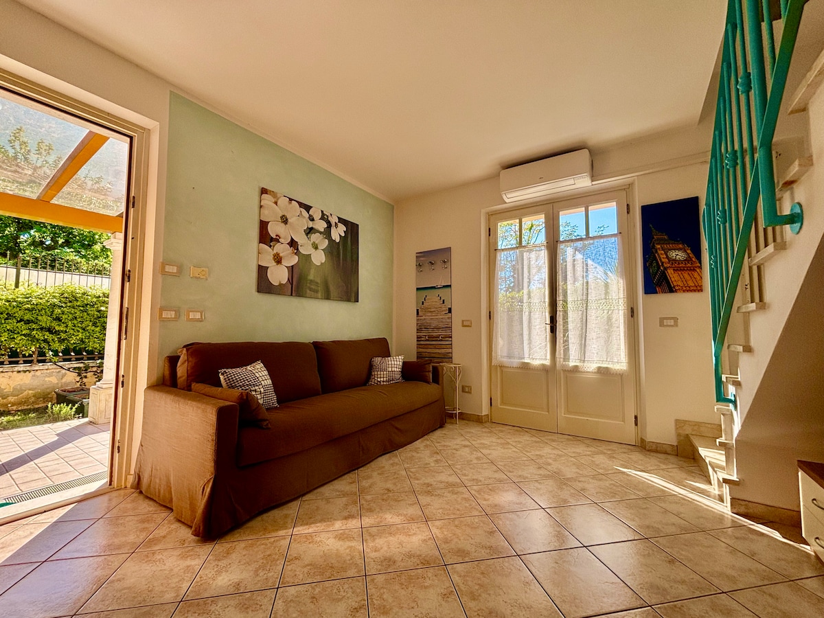 A spacious living area is presented with a brown sofa and decorative artwork on the walls. Natural light filters in through large windows, illuminating the tiled floor. A door provides access to an outdoor space, while a staircase leads to the upper level.