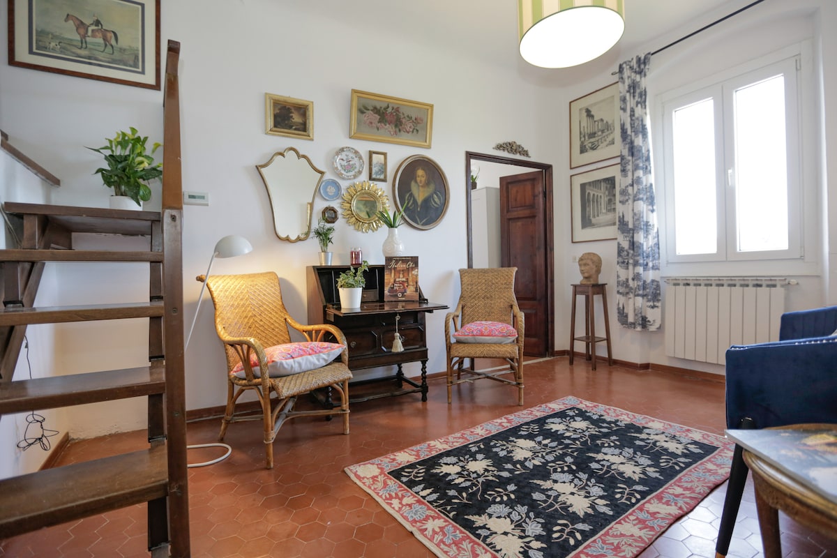 A cozy living area is presented, featuring two armchairs with floral upholstery, a small circular table, and a decorative mirror. A staircase leads to an upper level. The walls are adorned with framed artwork, and a large area rug adds warmth to the terracotta flooring.