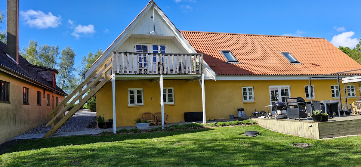 A yellow two-story building is surrounded by greenery, featuring a wooden staircase leading to a balcony. Large windows allow for ample natural light, while patio furniture is arranged on a deck in the foreground, inviting outdoor relaxation.