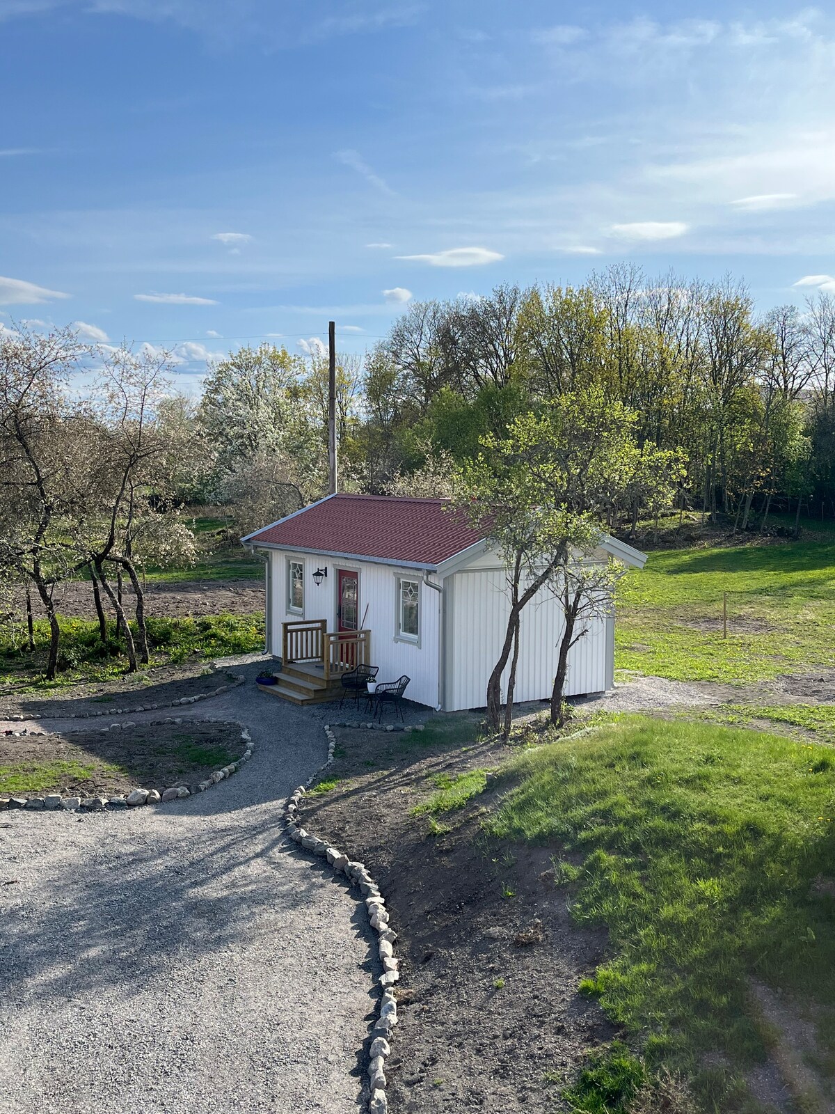 A newly built cabin is set in a serene countryside environment, featuring a red-roofed exterior and white wooden walls. A winding gravel pathway leads to the entrance, bordered by lush greenery and trees. Clear skies can be seen overhead, enhancing the peaceful setting.