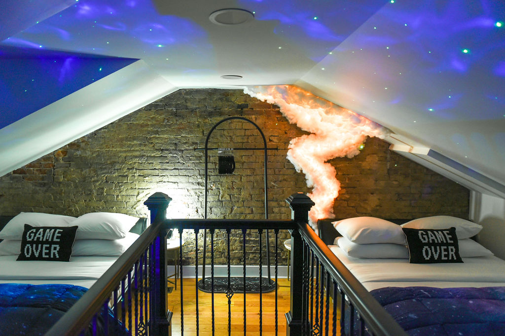 A cozy loft-style bedroom features two full beds under a glowing ceiling, simulating a galaxy. Each bed is adorned with black pillows inscribed with 'GAME OVER.' Exposed brick walls and soft lighting enhance the playful atmosphere of the space.