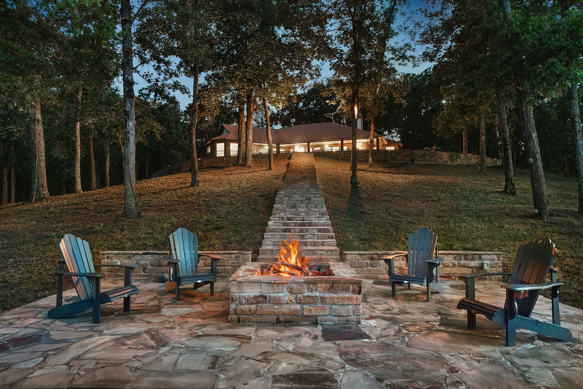 A stone fire pit is surrounded by four adirondack chairs on a spacious patio, offering a cozy gathering space. A gently sloping pathway leads to the well-lit house in the background, framed by tall trees, creating a serene outdoor setting.