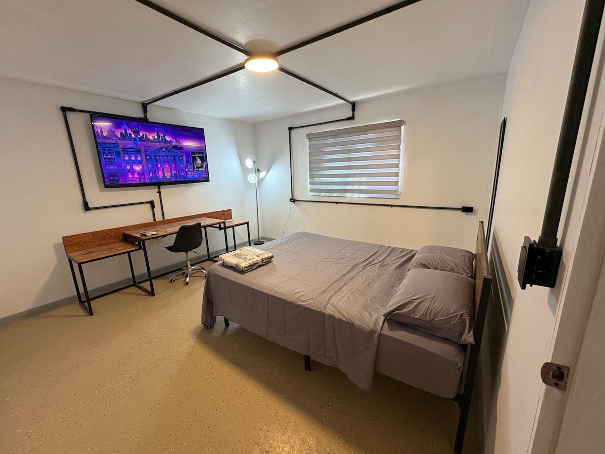 An industrial-style bedroom features a spacious bed with a neutral-colored duvet. A flat-screen TV is mounted on the wall, and a wooden desk with a chair is positioned nearby. Natural light enters through a window with light-filtering blinds.