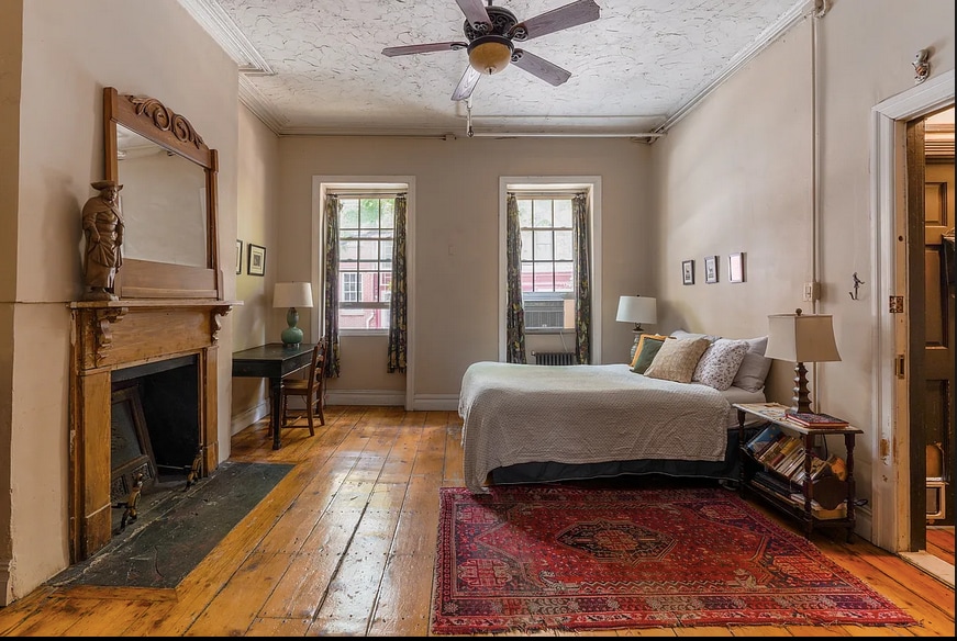 A spacious parlor apartment features original wide-plank oak floors and twelve-foot ceilings. The room includes a cozy bed with layered bedding, a decorative area rug, and a wooden fireplace mantel. Tall windows provide natural light, while a small dining table is positioned in the corner.