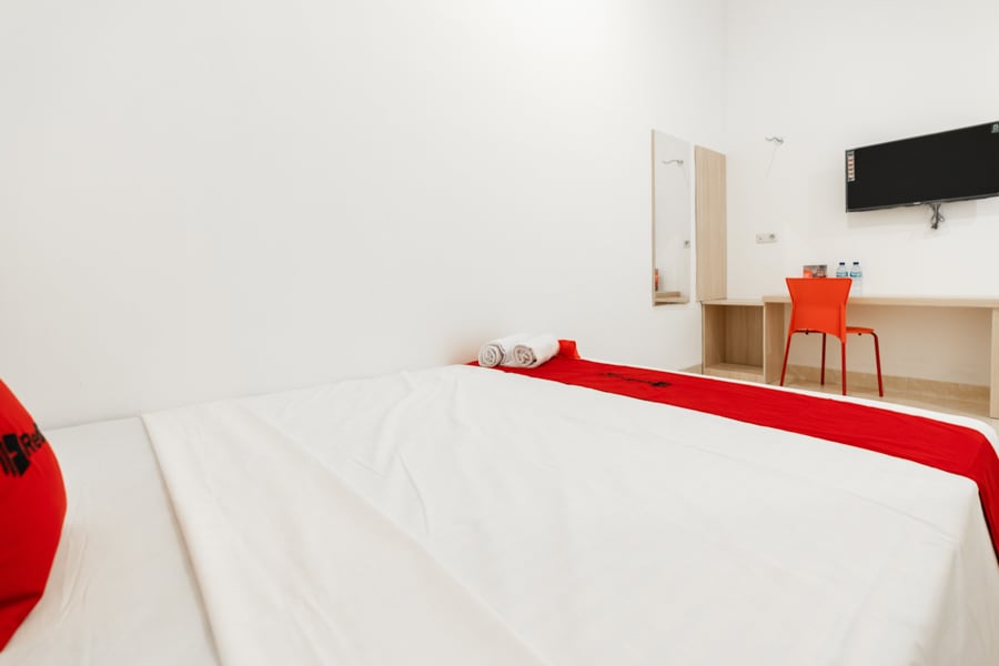 A spacious room is presented with a large, neatly made bed covered in white linens and a red accent blanket. A mirror is mounted on one wall, and a small work desk is paired with a bright red chair. Bottled water is visible on the desk.