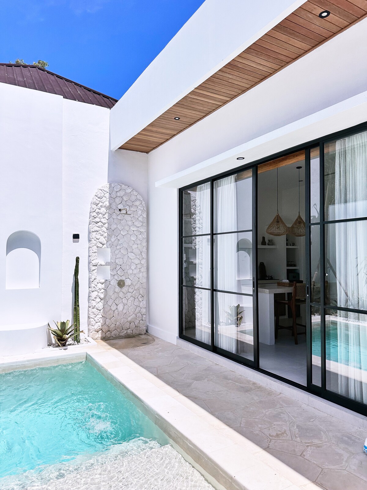A sunlit area is visible where a private pool meets a spacious deck. Glass sliding doors open to an elegantly designed indoor space, framed by soft curtains. Natural textures are evident in the exterior stonework, complemented by gentle greenery along the pool's edge.
