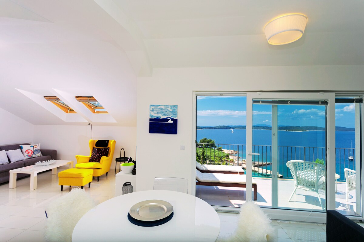 A modern living space features large sliding glass doors that provide a panoramic sea view. Natural light floods the room through skylights and windows. A round dining table is surrounded by white chairs, while a stylish yellow armchair adds a pop of color beside a cozy sofa.