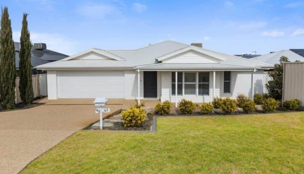 A single-story house featuring a light-colored exterior and a covered front porch. The neatly manicured lawn is complemented by low-maintenance shrubs along the pathway. A spacious driveway leads to a garage with a flat roof, while tall trees provide privacy on one side.