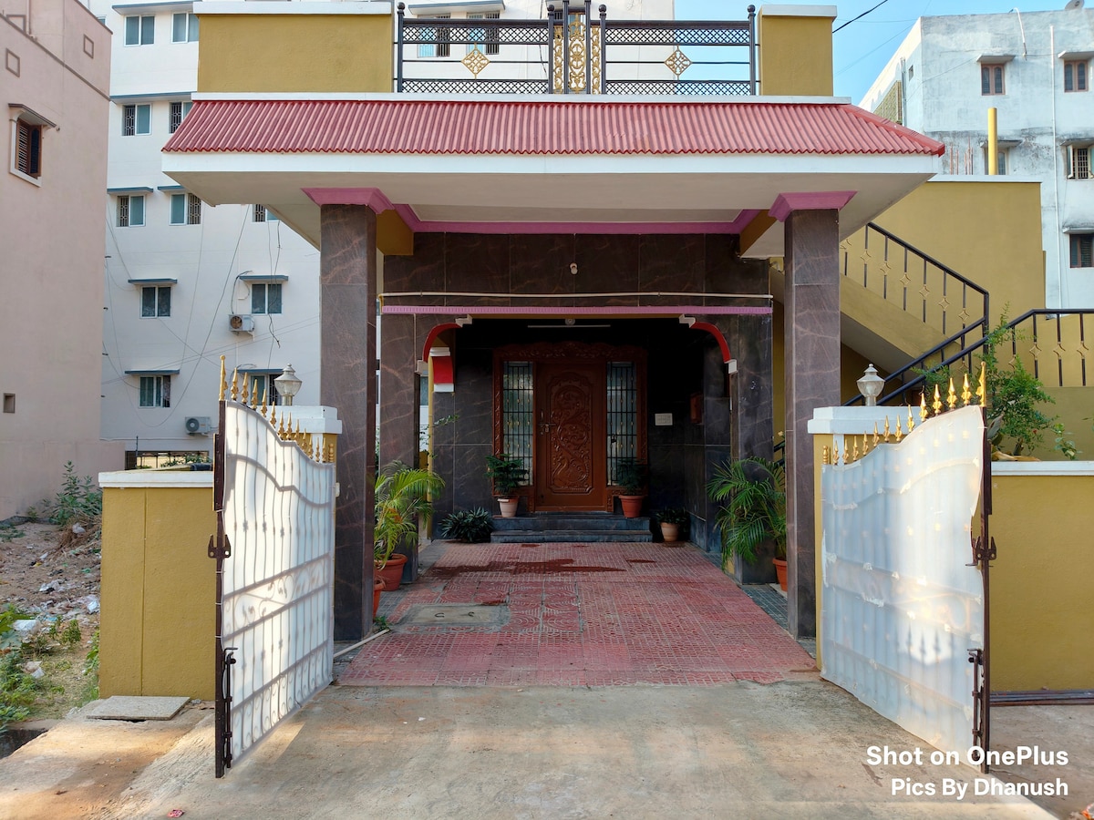 The entrance showcases a well-maintained facade with a decorative gate and a paved pathway leading to the main door. Potted plants line the entrance, contributing to the welcoming appearance of the home. The porch is sheltered by a sloped roof.