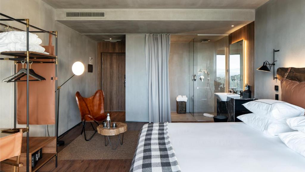 A modern hotel room is presented with a spacious bed adorned with multiple pillows. A lounge chair sits beside a wooden side table, and a stylish clothing rack displays folded towels. A glass-enclosed bathroom area features a clear view of the shower and sink.