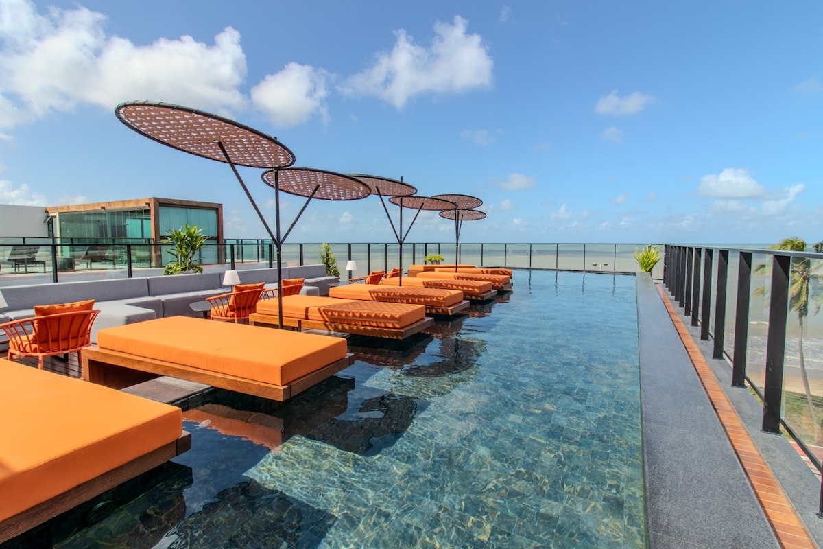A rooftop pool area is featured, surrounded by comfortable lounge chairs with orange cushions. Parasol shades provide sun protection, and a clear view of the ocean and sky creates a serene atmosphere. The well-maintained water reflects the surrounding environment, enhancing the relaxing setting.