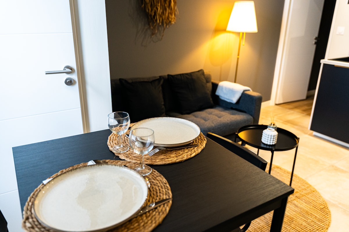 A dining table is set with two plates and glasses, positioned next to a cozy gray sofa. Soft lighting is provided by a standing lamp, while a small black table complements the space. The overall decor creates a modern and inviting atmosphere.