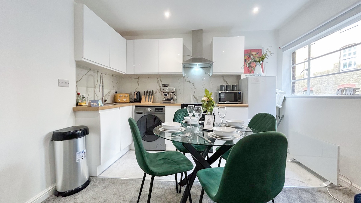The kitchen area features a modern design with white cabinetry and sleek marble accents. A round glass dining table with green chairs is set for a meal. Essential appliances and a coffee maker are visible, providing a functional and inviting space.