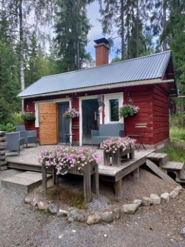 A rustic red wooden cabin is surrounded by tall trees, featuring a welcoming wooden door and flower boxes adorned with vibrant flowers. A spacious deck includes two armchairs and seating areas, inviting relaxation in a serene natural setting.