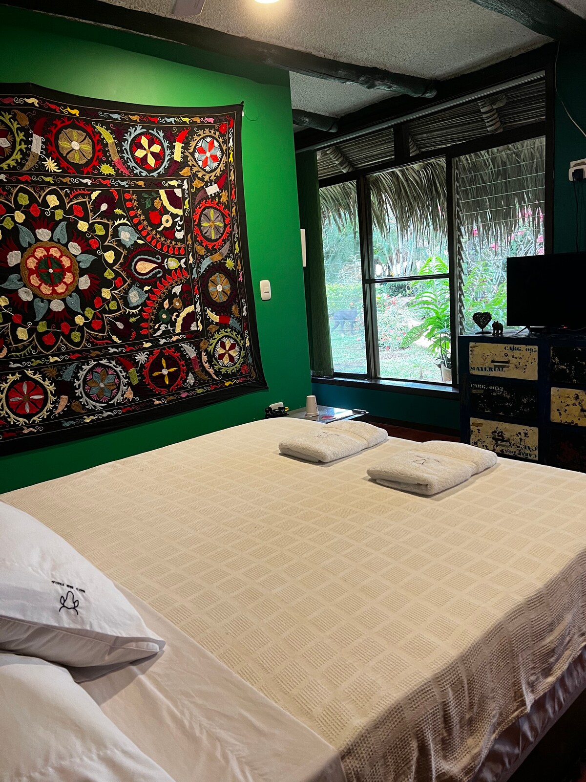 A cozy sleeping area features a spacious bed with neatly arranged towels. A richly patterned tapestry adorns the green wall above the bed. Large windows provide natural light and a view of lush greenery. A television is positioned on a wooden dresser.