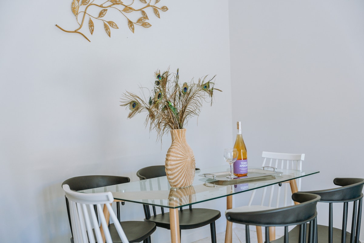 A glass dining table is surrounded by five chairs in contrasting black and white colors. A decorative vase filled with peacock feathers is positioned at the center, accompanied by a bottle of white wine and a glass, creating an elegant dining setting.