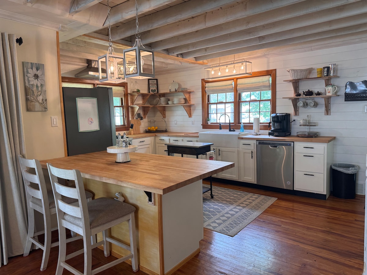 A modern kitchen space is highlighted by a central wooden island with seating. Open shelving displays elegant dishware, and warm wood tones complement the sleek appliances. Large windows allow natural light to fill the room, enhancing the inviting atmosphere and showcasing the rustic beams above.