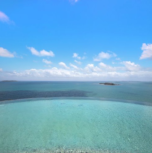 A serene coastal view captures the horizon where the turquoise waters meet a clear blue sky. Gentle waves create ripples across the sea, while distant islands are visible against the horizon, enhancing the tranquil atmosphere of this idyllic setting.