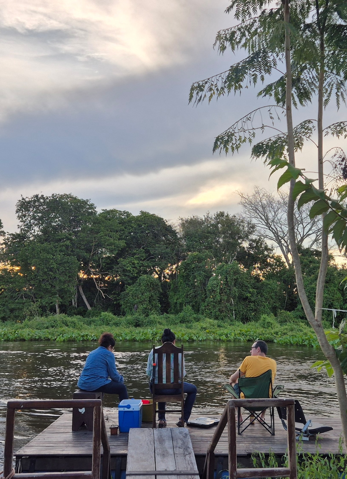 Two individuals are seated on a wooden dock facing a river, surrounded by lush greenery. A cooler is placed beside one person, while the serene water reflects the evening sky with soft hues. Trees line the opposite bank, enhancing the natural ambiance.