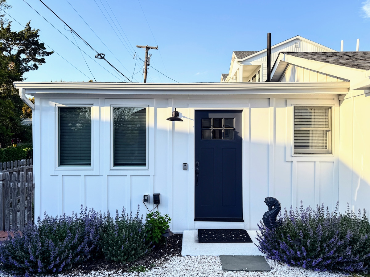 The exterior of the cottage features a welcoming entrance with a navy blue front door, flanked by two windows with grid patterns. Lush lavender plants line the pathway, leading to the door, while a decorative element sits beside the entrance.