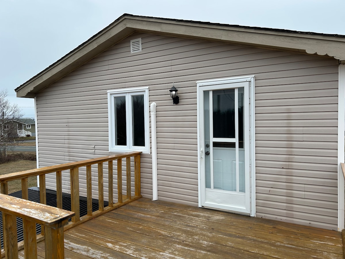 The exterior of a house is shown, featuring light-colored siding and a wooden deck with a railing. A white door is positioned at the center, flanked by two windows. The surrounding area is open, indicating a tranquil setting.