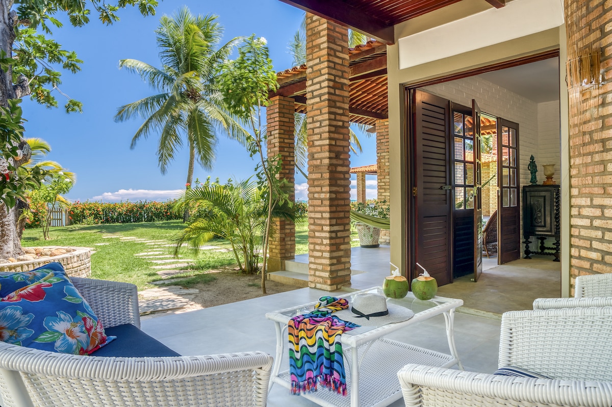 A bright outdoor seating area is presented, featuring white wicker furniture and colorful cushions. Lush greenery and palm trees provide a tropical ambiance. The view extends towards a sunny landscape, while the entrance into the spacious interior is framed by wooden doors, inviting relaxation and connection with nature.