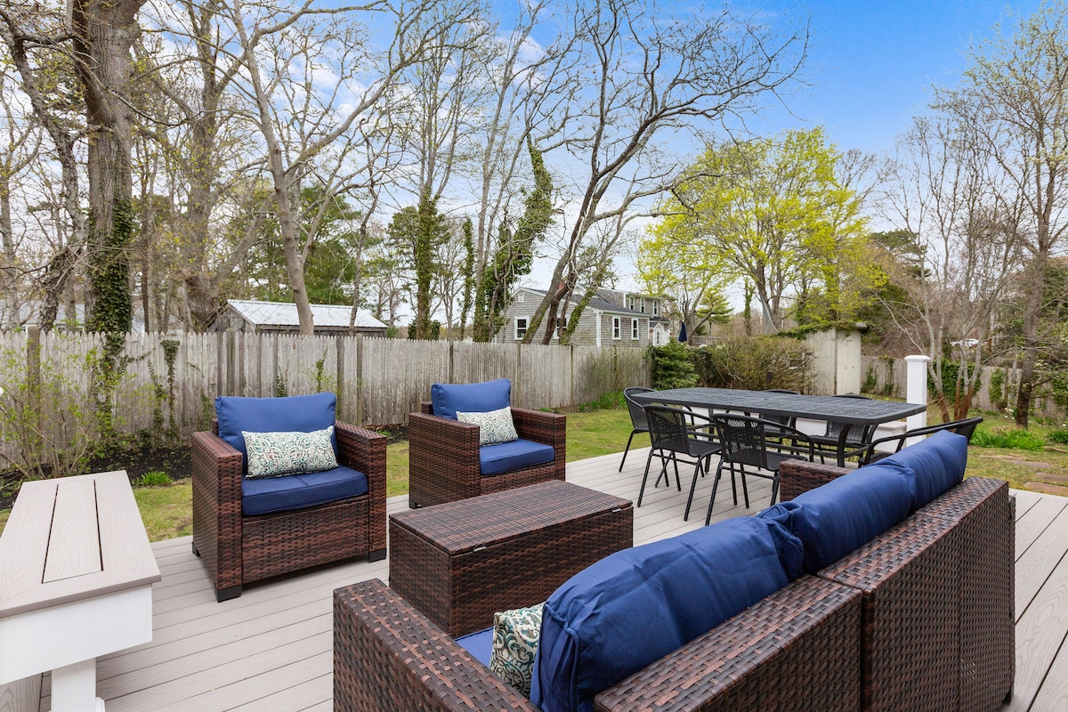 An outdoor deck is showcased, furnished with a seating area of dark wicker furniture and blue cushions. A dining table for six is positioned adjacent to the seating area, surrounded by a spacious backyard featuring trees and a wooden fence.