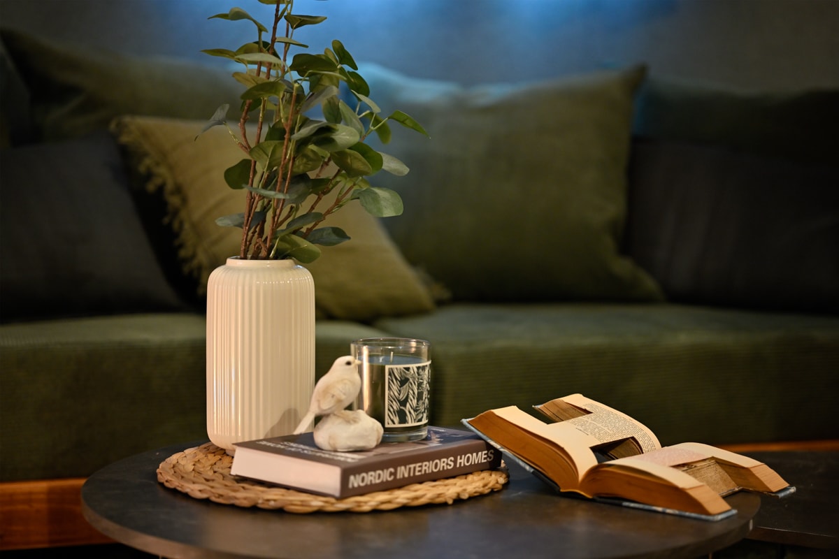 A cozy seating area features a green upholstered sofa adorned with plush pillows. A round coffee table holds a vase with leafy greenery, a candle, a small decor figurine, and an open book, creating an inviting atmosphere for relaxation.