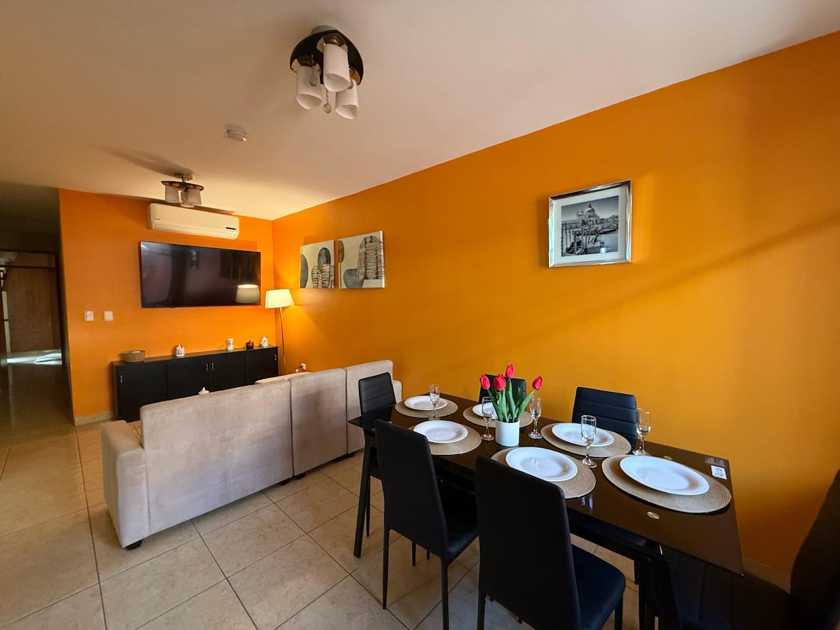 A bright living area features an orange accent wall and modern furnishings. A comfortable sofa is positioned against the wall, complemented by a dining table set with flowers. A TV and decorative wall art are displayed, enhancing the overall ambiance of the space.