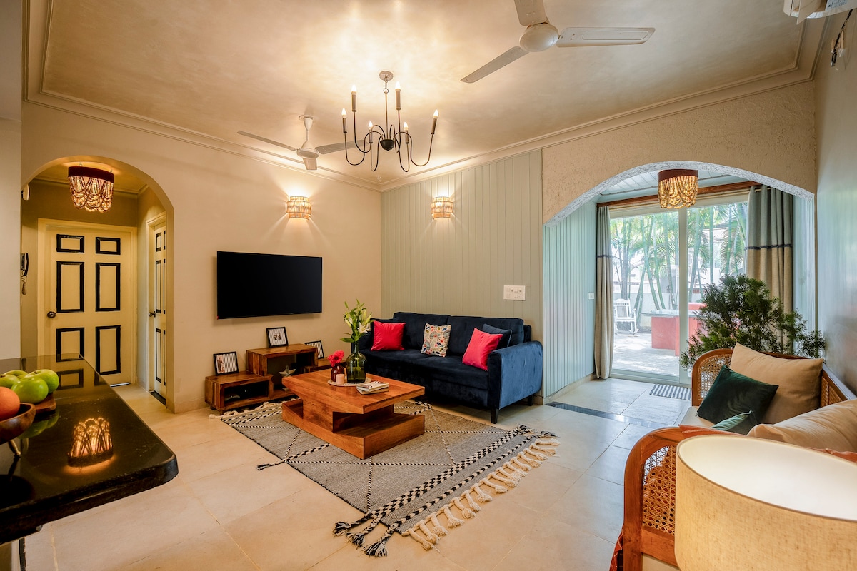 The living area features a comfortable blue sofa with patterned pillows and a wooden coffee table atop a decorative rug. Natural light enters through large glass doors that lead to the private garden. The space is complemented by a TV mounted on the wall and elegant light fixtures.