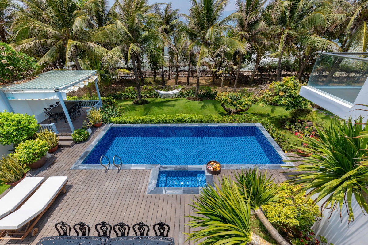 An outdoor area features a sparkling blue pool surrounded by vibrant greenery and palm trees. A wooden deck accommodates lounge chairs and a dining area, with a hammock visible in the background. Colorful plants add to the inviting garden atmosphere.