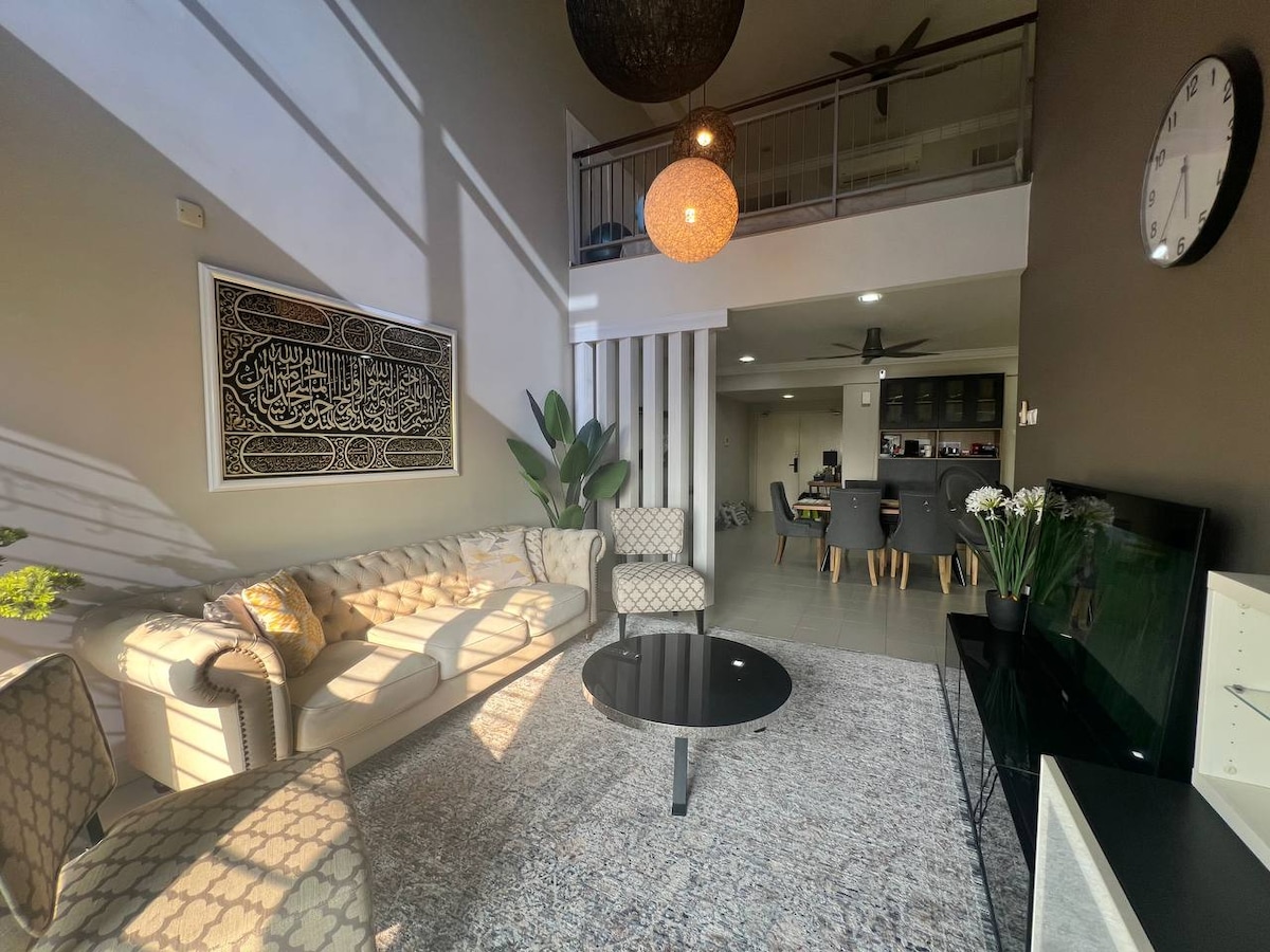 A stylish living area features a plush sofa complemented by patterned armchairs. A round black coffee table sits atop a soft area rug. Natural light streams in through large windows, while decorative plants and wall art enhance the space's modern design.