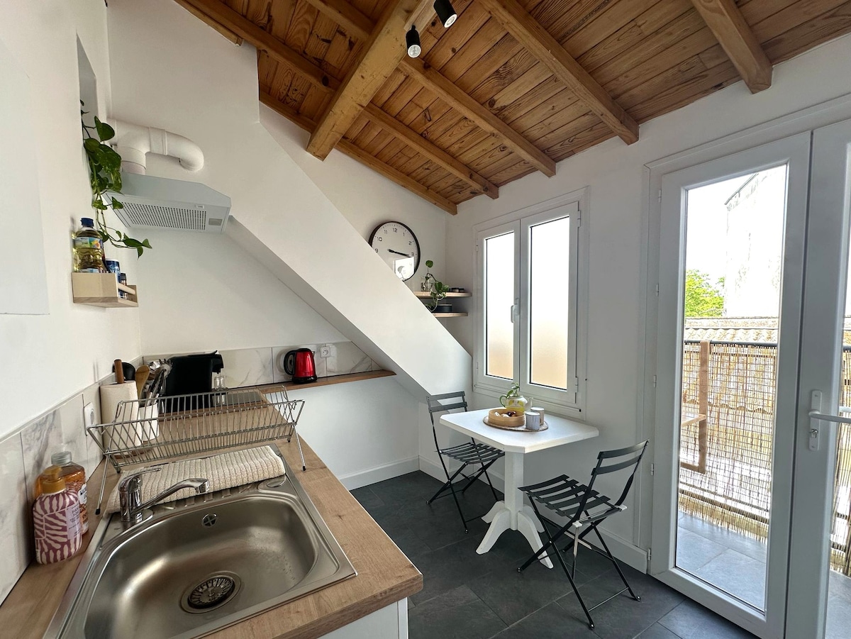 A well-lit kitchen area features a sink, wooden countertop, and stainless steel appliances. A small table with two metal chairs is placed near a set of glass doors that access the private balcony. The ceiling showcases wooden beams, adding depth to the space.