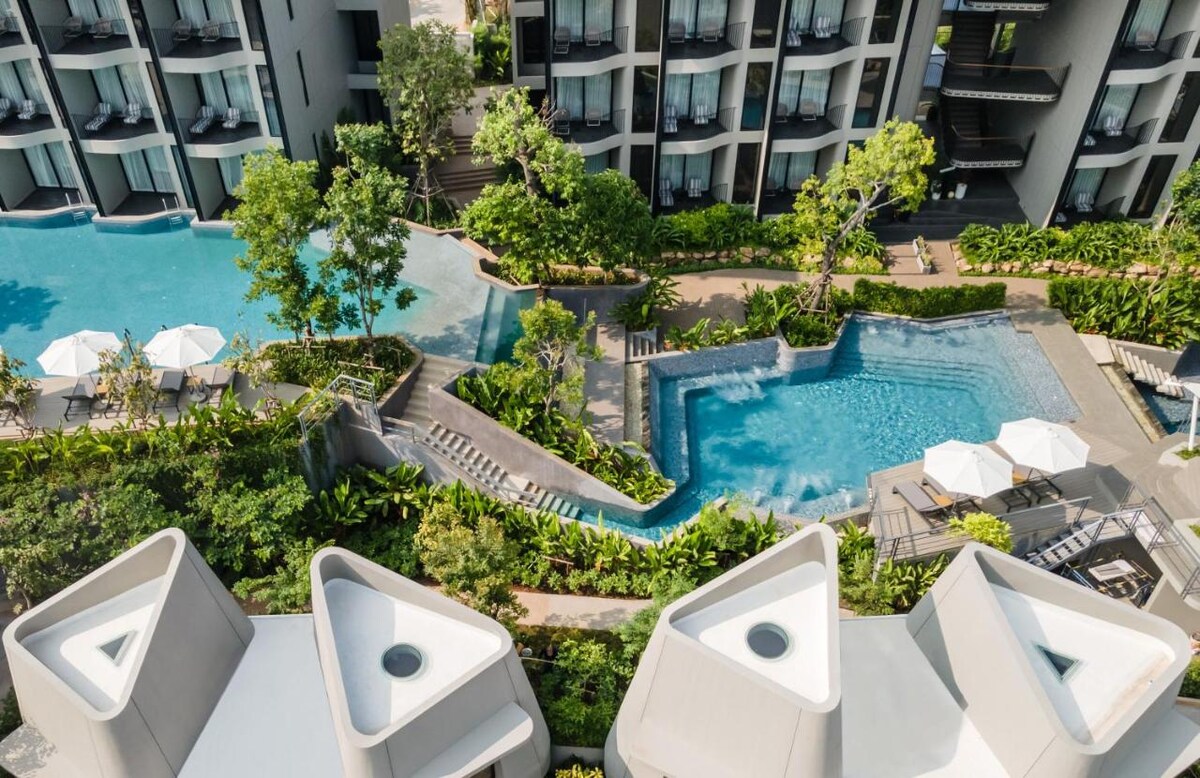 Aerial view of the property showcases multiple infinity swimming pools surrounded by lush greenery. Sun loungers with umbrellas line the poolside, while landscaped pathways lead to different areas. Modern buildings provide a backdrop to the serene outdoor spaces.