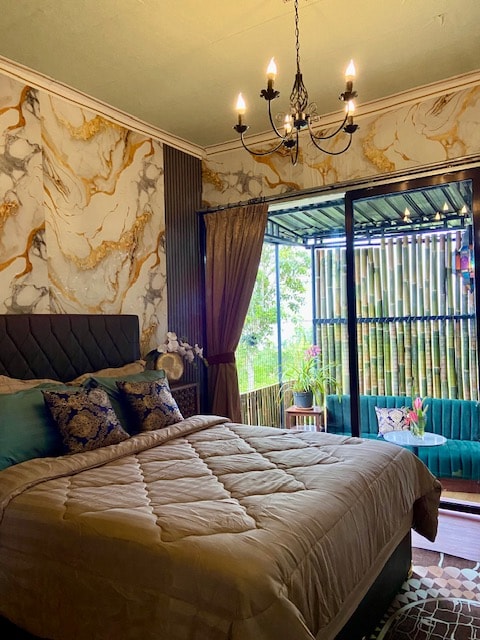 A bedroom features ornate marble-patterned walls and a spacious bed adorned with decorative pillows. Natural light filters through large sliding doors that open onto a small outdoor seating area, surrounded by greenery and bamboo, creating a serene atmosphere.