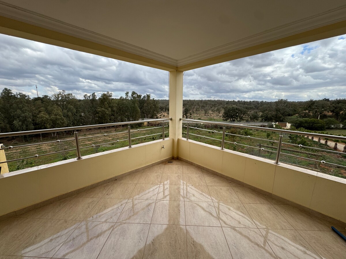 A spacious balcony offers panoramic views of lush greenery and open fields. The polished floor reflects natural light, creating a bright and airy atmosphere. Metal railings frame the view, enhancing the connection to the surrounding landscape, ideal for enjoying fresh air and tranquility.