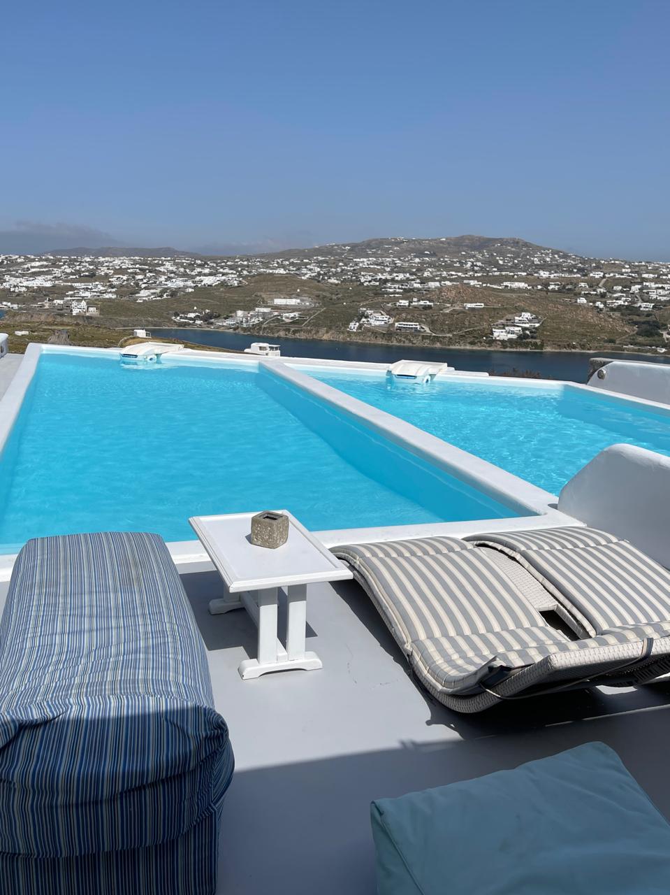 The rooftop area features two long, turquoise swimming pools side by side, with a scenic view of the surroundings. Comfortable lounge chairs are positioned nearby, accompanied by a small white table. The calm water reflects the clear blue sky above.
