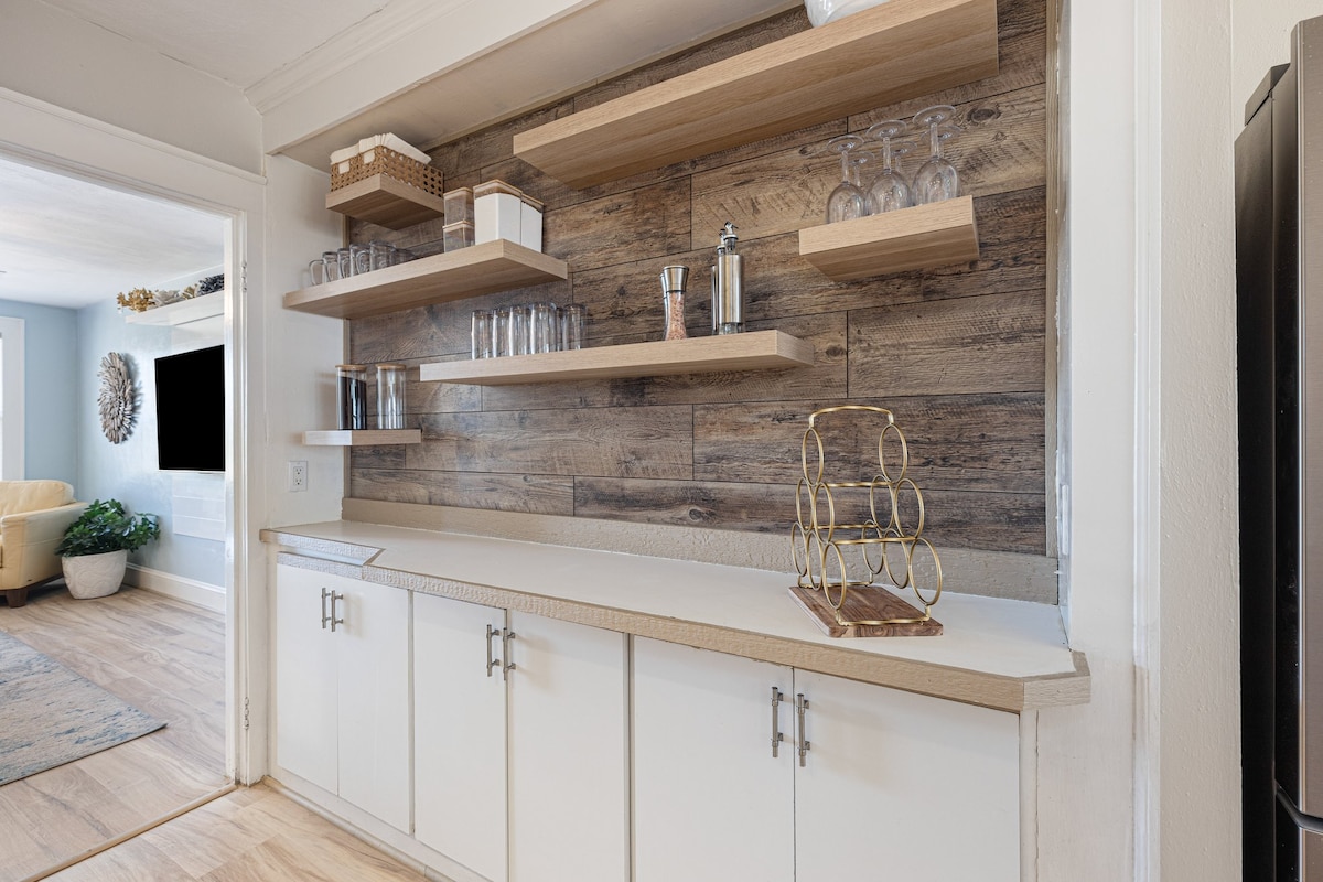 A kitchen area features open shelving displaying an array of glassware and a few decorative items. The countertop is smooth and uncluttered, complemented by white cabinetry with modern handles. Rustic wood paneling forms the backdrop, creating a warm and approachable space.