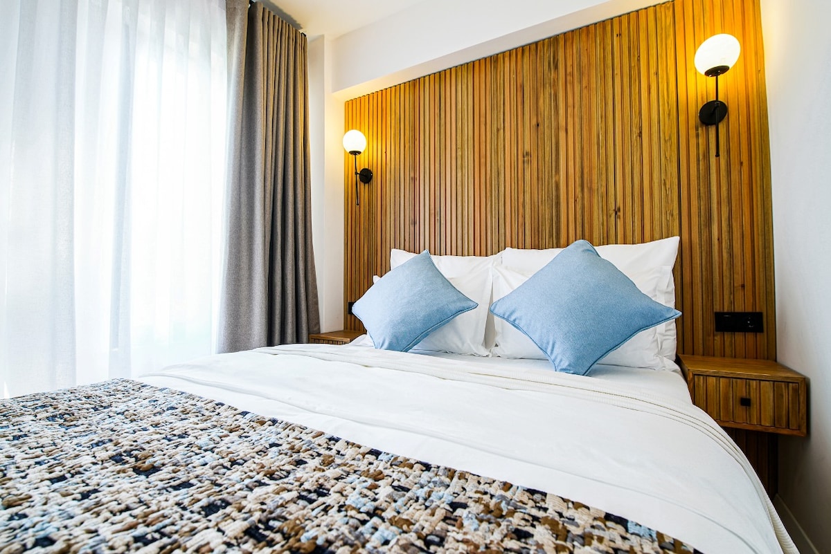 A comfortable double bed is adorned with two blue accent pillows. The wooden headboard adds a natural element to the space, complemented by wall-mounted lights. Sheer white curtains frame a window, allowing light to softly illuminate the room.