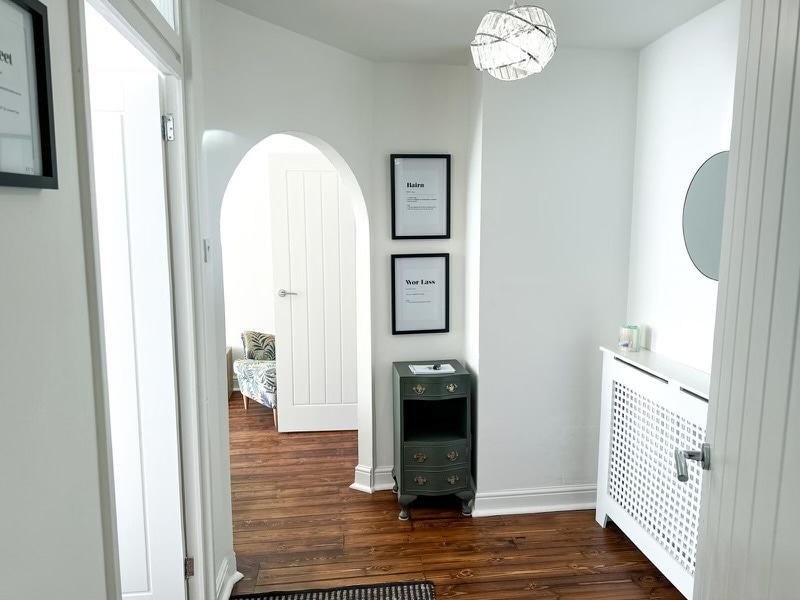 An entryway features light wooden flooring and bright white walls. A stylish light fixture hangs from the ceiling. A circular mirror is mounted on the wall, alongside two framed prints. A small green cabinet stands next to a radiator, providing functional storage.