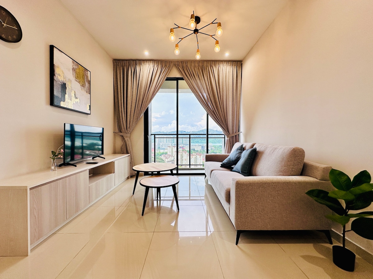 A modern living area features a comfortable gray sofa and two round coffee tables positioned on glossy tile flooring. Large windows, framed by light curtains, provide a view of the city landscape. A television is mounted on the wall, and a small potted plant adds a touch of greenery.