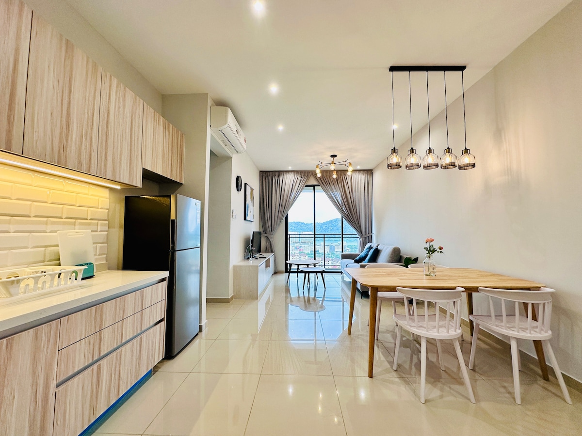 A modern and open living area features a well-equipped kitchen with light wood cabinetry and stainless steel appliances. A dining table for four is positioned next to large windows, allowing natural light to fill the space. Comfortable seating is arranged in a cozy corner.