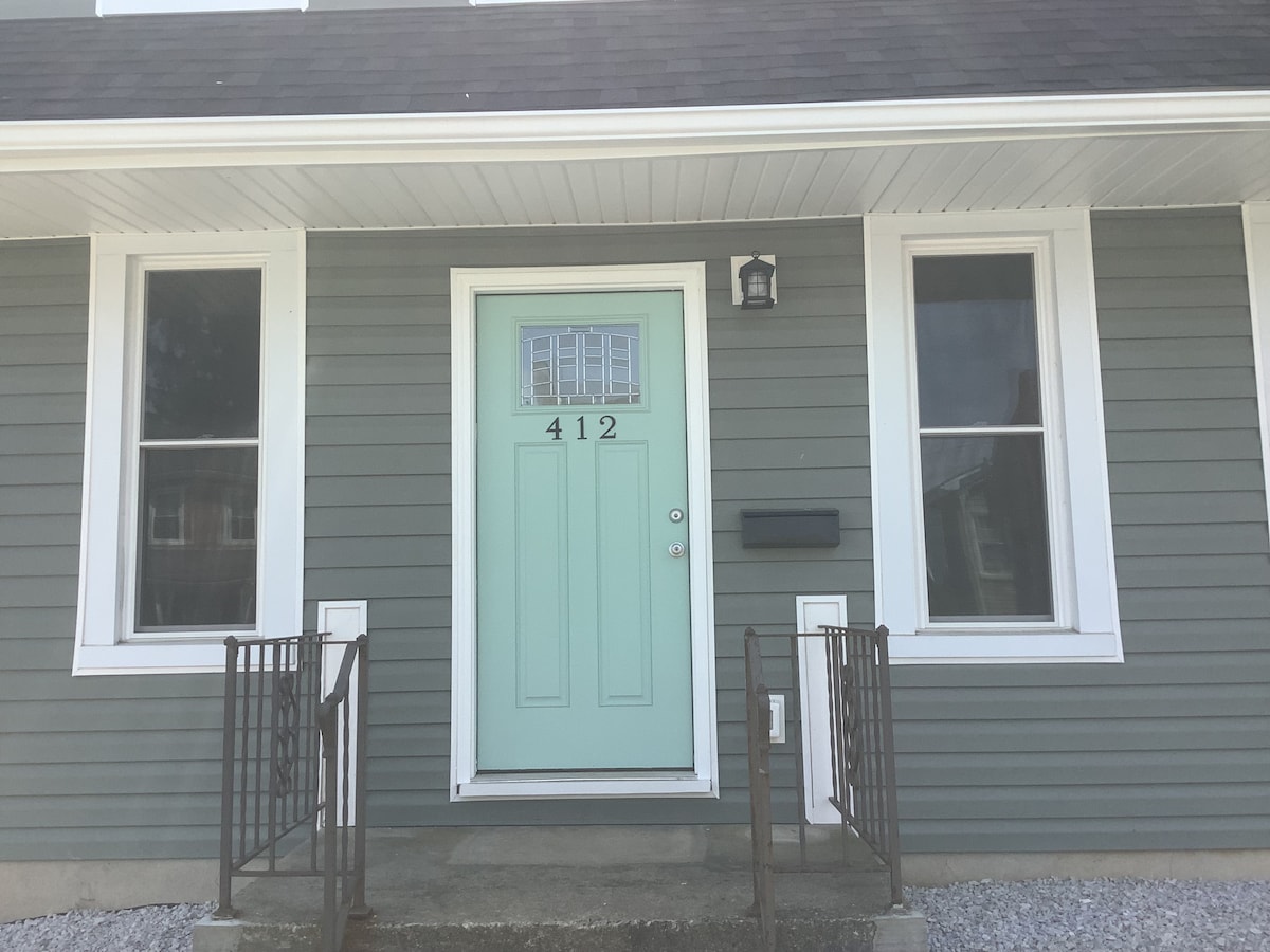 The entrance features a mint green door with glass accents, framed by large windows on either side. A concrete step leads up to the door, which is flanked by decorative metal railings. The gray exterior siding provides a modern touch to the home.