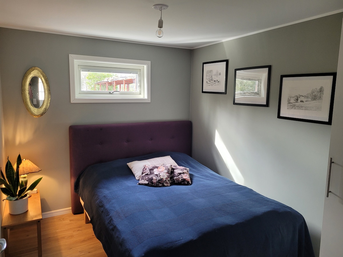 A cozy bedroom is presented, featuring a spacious bed with a dark blue coverlet. Framed black-and-white artwork hangs on the wall, complemented by a small bedside table with a lamp and a decorative plant. Natural light filters through a narrow window.