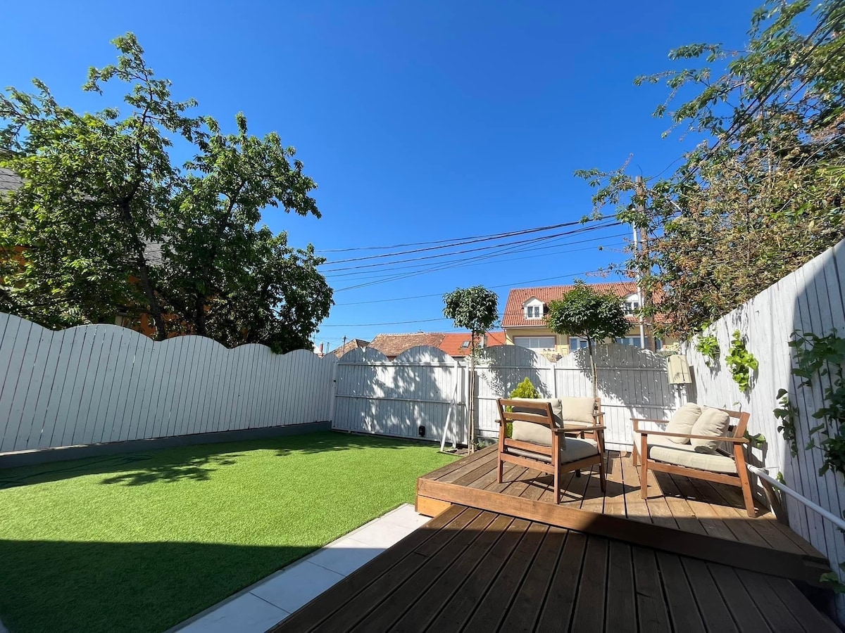 A private outdoor area features a well-maintained green lawn surrounded by a white fence. Comfortable seating made of wood is positioned on a wooden deck, providing a pleasant space to relax. Clear blue skies and a few trees add to the inviting ambiance.