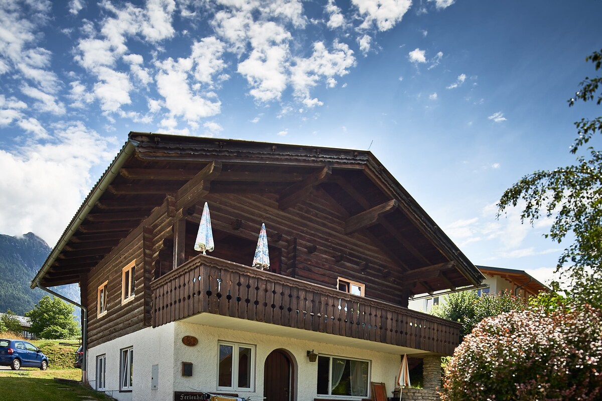 The exterior of a wooden chalet presents a charming alpine design, featuring a balcony adorned with two colorful umbrellas. Lush greenery surrounds the building, underlined by a bright blue sky dotted with clouds, highlighting the mountainous backdrop.