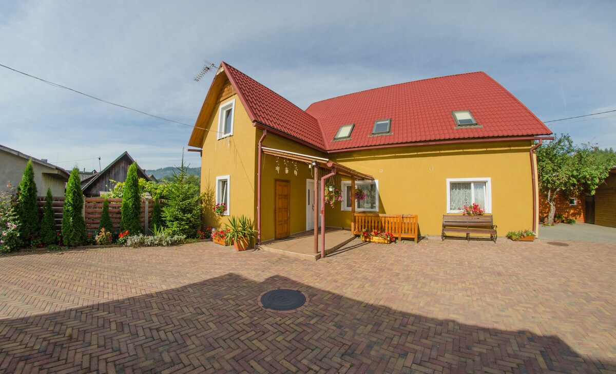 A charming yellow house is displayed with a red roof and white trim. The spacious patio is paved with brick and features colorful flower beds. Surrounding greenery adds to the warmth of the property, while a wooden bench offers a welcoming spot for relaxation.