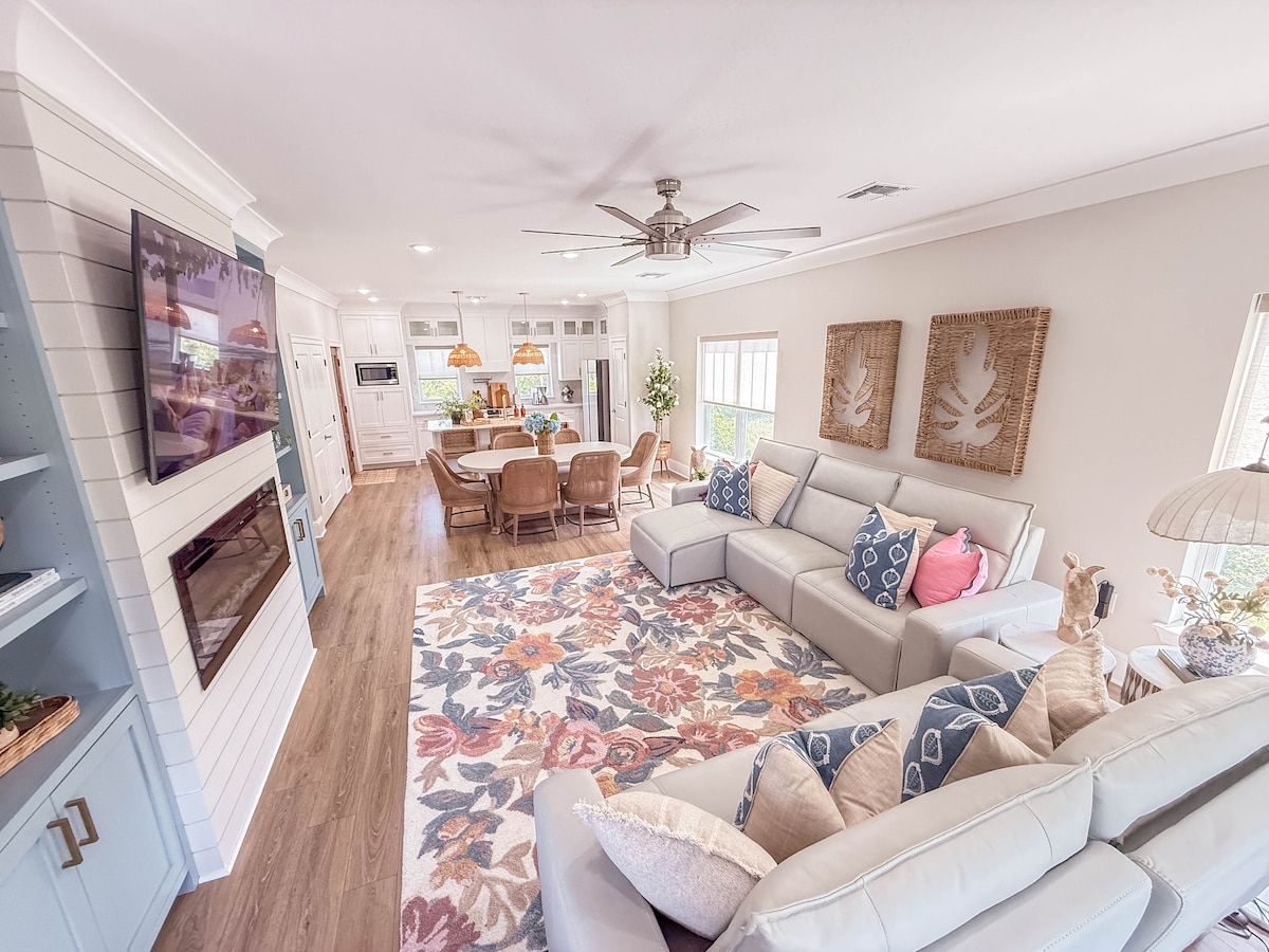 A spacious open-concept living area features a comfortable light-colored sectional sofa facing a mounted television and a decorative fireplace. A large area rug with floral patterns is laid on the floor. To the back, a dining area with a wooden table and wicker chairs is visible, along with a modern kitchen.